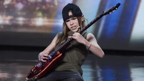 Britain's Got Talent/Freemantle/SYCO Olly playing a red electric guitar on his knees with a look of deep concentration. He is on stage, and wearing a black back to front baseball cap, a green vest top and black shorts. His hair is light brown and reaches beyond his shoulders.