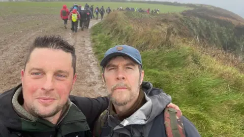 Maxwell Family Fund Craig Maxwell smiles and has an arm around comedian Rhod Gilbert's shoulder. The pair are in the middle of a muddy path with a group of hikers in the background.