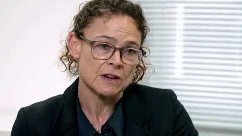 A woman with curly hair and glasses speaks to the camera. She is wearing a balck jacket, and behind her is a white wall and blinds