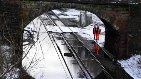 Two workmen in orange high-vis gear survey a flooded railway track passing under a bridge, surrounded by snow.