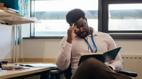 Getty Images A teacher sits in a school office, reading from an open folder with his hand to his head. The desk nearby is scattered with papers and office supplies, and shelves above hold neatly arranged binders. There are windows behind the teacher letting in light.