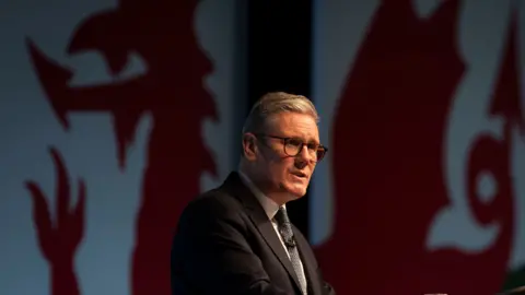 A man with white hair, glasses, and wearing a navy suit makes a speech in front of a big Welsh dragon flag.