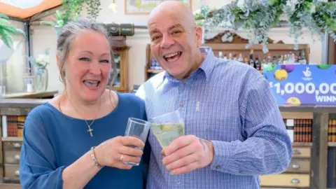 PA Media Caroline (left) and Terry Gillings celebrating their win with a drink. Caroline has grey hair, tied back, and is wearing a blue top. Her husband is bald and wearing a blue checked shirt. Behind them is a sign that reads "£1,000,000 winner".