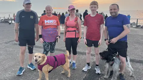 Zoie O'Brien/BBC A group of two men, two women and a teenage boy, all in running gear of shirts and t-shirts stand side by side and smile at the camera. Standing in front of them are two dogs - a black and white mixed breed and a blond labrador. Behind them is a promenade and sea. 