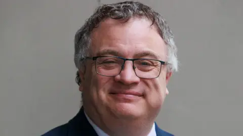 Stephen Farry, a man with grey-black hair and black-rimmed glasses, smiles at the camera