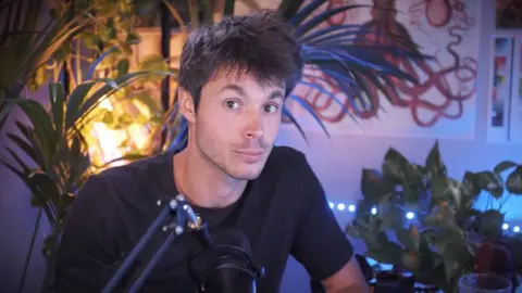 Léo Grasset Image of a YouTuber sitting in a dimly lit room with a microphone in front of him and pot plants behind him. He is looking straight at the camera and has his eyebrows raised.