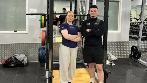 Hutton Moor Lesuire Centre Phoebe Pothecary standing in front of a weight rack at the gym with her arms folded, beside another man