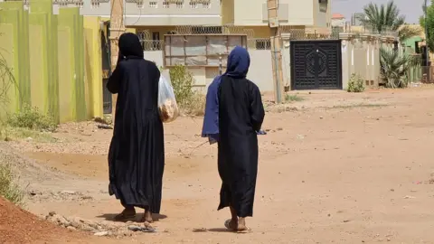 Getty Images Two women in black cloaks walking towards residential buildings, their backs to the camera. One woman is carrying a bag full of bread.