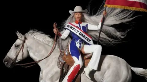 Beyonce / Parkwood Album art for Beyoncé's album, Cowboy Carter. Beyoncé is a 42-year-old black woman. She's styled in a red, white and blue patent cowboy-themed outfit including a cowboy hat, chaps and a buttoned up shirt. She's pictured sitting side-saddle on a white horse, her long silver hair flowing behind her. In her right hand, she holds the horse's reigns and in her left she holds aloft the American flag.