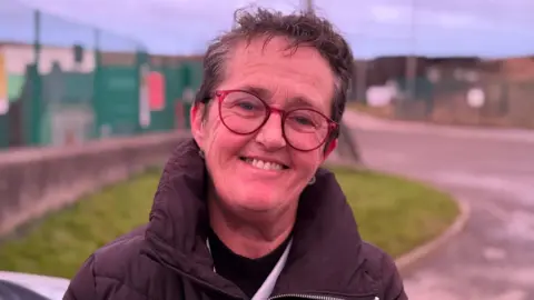 Alyson stands in front of the landfill site. She smiles at the camera. She has short, dark hair. She wears red, round glasses and a dark puffer jacket.