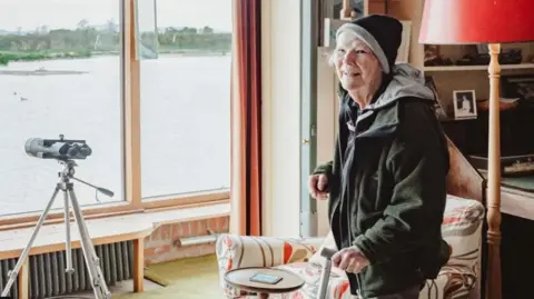 WWT Bettie in outdoor gear and woollen hat, smiling to camera, standing in a room overlooking a lake with a telescope in the window