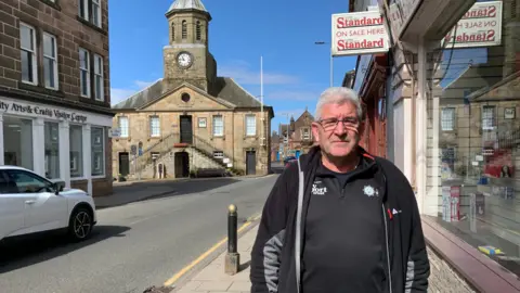 A man in a black top and jacket stands outside a shop with a historic Tolbooth building in the background