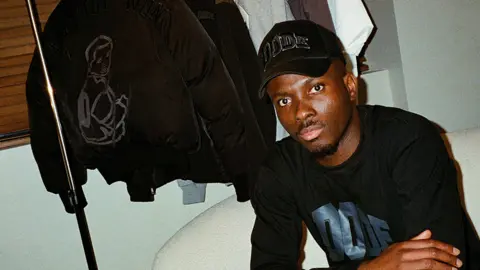 Dimeji Ode sitting on a sofa with a rack of his Dode clothing in the back. He wears a black baseball cap with the word "Dode" on it and wears a black top with "Dode" written on it in blue. His has black trousers with a grey vertical stripe. He looks into the camera and has a short goatee beard and moustache.