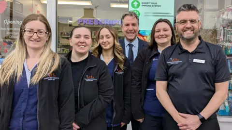 Dan Norris West of England Mayor Dan Norris, wearing a dark grey suit with blue shirt and dark blue tie, stands at the back of a group of people. The rest of the group are staff members at the Cadbury Heath Pharmacy and Health Clinic and are wearing branded black fleeces. They are a mixture of men and women