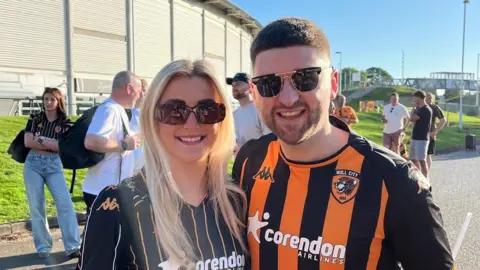 Richard Madden/BBC A man with short brown hair and a woman with long blonde hair smile for the camera. Both are wearing Hull City shirts. Behind them is a football stadium.