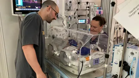 A man in dark grey hospital tunic looking at an baby in an incubator with a nurse covering the infant's head 