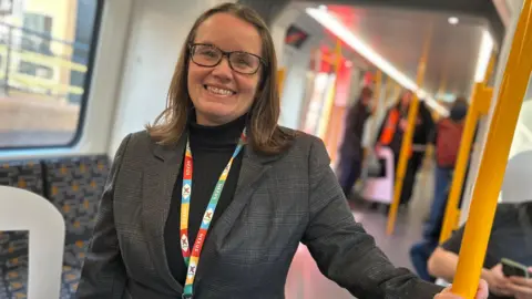 LDRS Nexus managing director Cathy Massarella smiling as she hold a yellow hand rail while on board the first new Tyne and Wear Metro in December 2024. She is wearing grey suit jacket over a black turtle neck top. She has glasses and a colorful lanyard. Her brown hair curls at the ends and reaches her shoulders. Behind her two men are sitting and looking in their phones. Other passengers are standing further back in the train. 