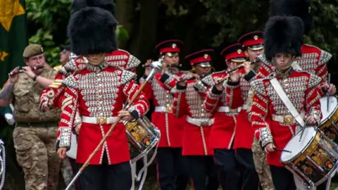 A military parade with soldiers in red ceremonial uniform with metal buttons and large fur hats or caps. They are holding or playing drums or wind instruments. 