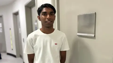 BBC/ Kate Bradbrook 18 year old man with a white t-shirt stands in a white corridor smiling at the camera