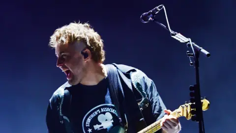 Sam Fender playing guitar next to a microphone on a stand. He is grinning and wearing a navy t-shirt.