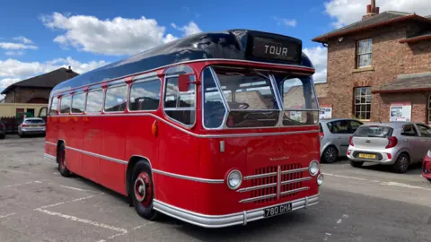 An old-school red bus with a black route parked on a car park. The bus is surrounded by other cars and brick buildings.