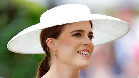 A close up shot of Princess Eugenie, wearing her hair in a low ponytail with a wide brimmed white hat. She is smiling and looking off to the right.
