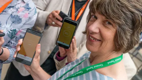 Bluestack Media The Bishop of Gloucester, Rt Revd Rachel Treweek, holding a phone showing the app