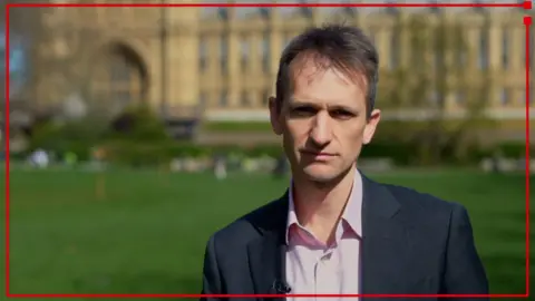 BBC reporter wearing a suit standing outside Houses of Parliament.