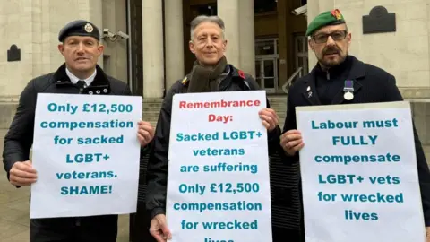 Adrian Radford-Shute Three men, two of whom are armed forces veterans standing in a row holding placards that have messages about £12500 compensation for LGBT+ veterans not being enough. The veteran on the right is Adrian Radford-Shute.