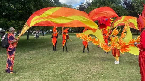 Carroll Weston/BBC People wearing red and orange outfits, posing as flames. They are waving large red and yellow banners in a park.