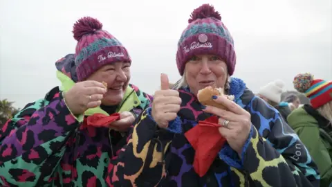 Two women smile and laugh at the camera wearing bright, multi-coloured leopard print dry robs both wearing knitted purple and blue beanies. They are eating a bap and holding red napkins in their hands.