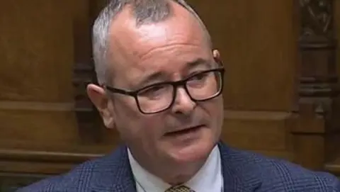 Lee Barron wearing a blue suit, white shirt and gold tie while speaking in the House of Commons.