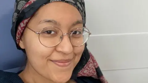 Dipika Saggi Dipika is looking at the camera, wearing a headband during her treatment. She has glasses on and is smiling. The background is blue and light grey.