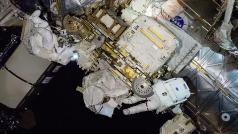 NASA Butch and Suni floated amid highly technical equipment outside the ISS, with blank space suits. You cannot say who, as one, with back to the camera, the other has its feet in the air. 