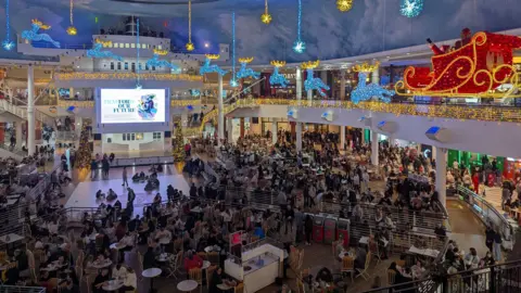 Dan Thomas Una vista hacia abajo de un área de patio de comidas abarrotada de gente, con decoraciones navideñas que incluyen un trineo de Papá Noel y renos suspendidos del techo.