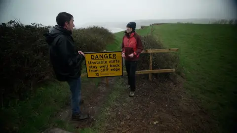 BBC /Kevin Church Lorna Sherriff from South West Coast Path Association stands with the reporter, Justin Rowlatt, by a National Trust yellow sign saying danger - unstable cliffs, stay well back from cliff edge. Both look west and the sky and the sea in the background are grey. The path is closed with a wooden gate just behind where they are standing. A cliff edge is seen a few meters after the wooden gate. 