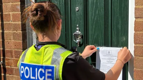 A female police office with brown hair in a bun. She is standing in front of a green front door with brick walls. She is sticking an A4 white piece of paper on the door with details of the closure.  