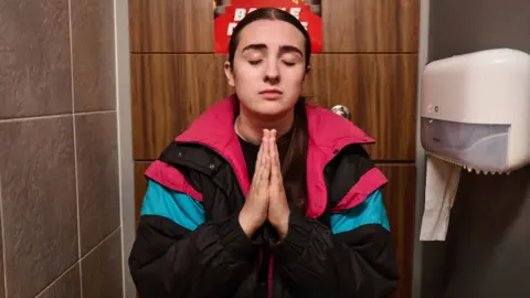 A young woman wears a shell suit and prays with her eyes closed in a toilet cubicle.