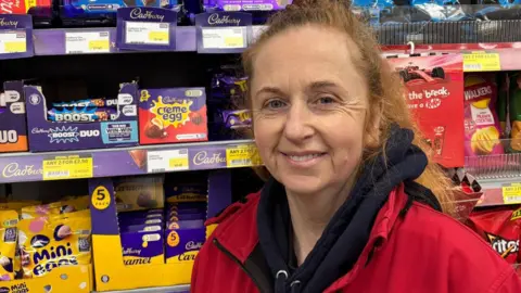 Fiona Avenal Malone Fiona Avenal Malone wearing a red fleece over a dark blue hoody standing in her shop in front of shelves of chocolate items