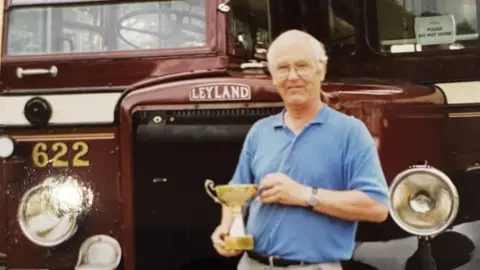 A man holds a gold trophy while standing in front of a red bus. The bus has a Leyland logo above the grill.