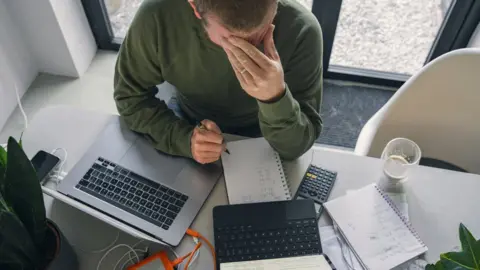 Getty Images A man is seen with his head in his hand while going through his bills at a table