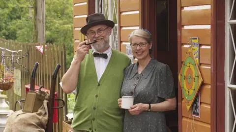 A smiling man and woman in vintage 1940s-style clothing standing outside a  small shop. The man is smoking a pipe and is wearing a hat and green vest. The woman is wearing a partnered dress and is holding a mug. Around them are baskets, crates, and retro goods.