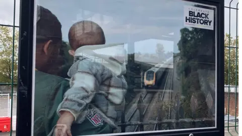 A photo of a man carrying his child, looking out across the railway