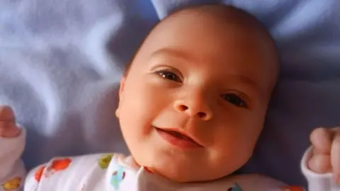 Sheree Black Baby Cameron. He is lying on a blanket with his closed hand by his head. He is wearing a baby grow with little characters on it.