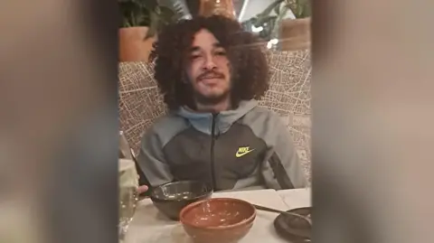An image of a man with dark, curly, shoulder-length hair. He is wearing a grey Nike zip top and is sat at a table with some empty bowls on. He is looking at the camera. 