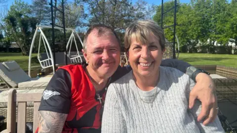 A man in a red and black T-shirt with tattoos on his arm hugs a woman in a grey jumper while both smile at the camera. In the background there is a grey outdoor seating setting and green grass. 