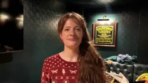 Alyssa Heath, a young woman wearing a red Christmas jumper and with red hair, stands in what appears to be a pub with green walls 