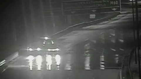 Black and white image shows flooding on surface of the M60 which has led to the motorway being closed so there is no traffic except for one vehicle on the hard shoulder. 