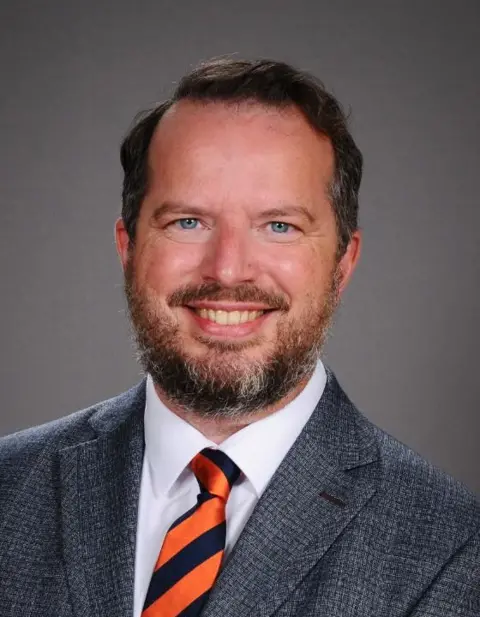 States of Guernsey The image shows Anthony Griffin smiling at the camera. He has short dark hair and a beard. He's wearing a grey blazer and a black and orange striped tie.