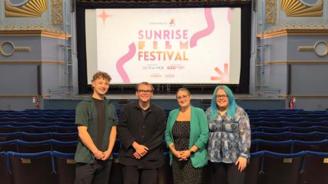Four people are standing beside each other in front of theatre stage showing sign for Sunrise Film Festival